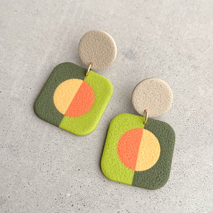 A pair of square drop earrings with a retro color-blocked design in green, yellow, and orange, hanging from textured beige circular studs on a light gray concrete surface.